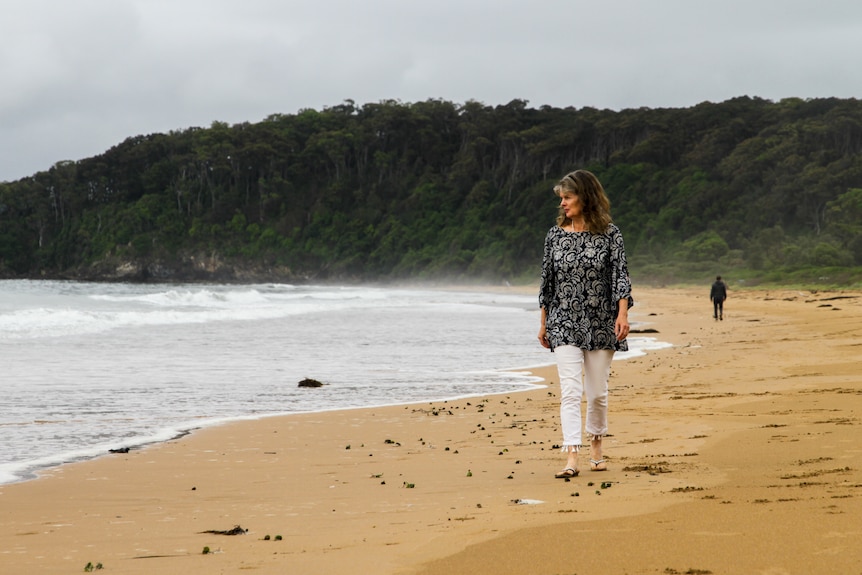 A woman in black and white top, white pants, walks along a beach while looking out to sea, green hills behind.