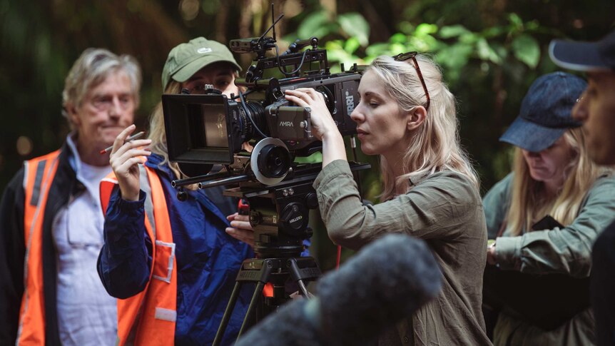 On set in rainforest, woman looks down lens of camera with other crew around her