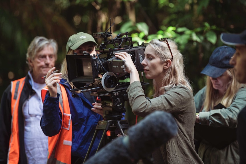 On set in rainforest, woman looks down lens of camera with other crew around her