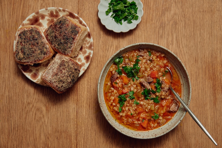 A bowl of red-cloured soup with grains and chunks of lamb, next to plates with bread rolls and parsley.