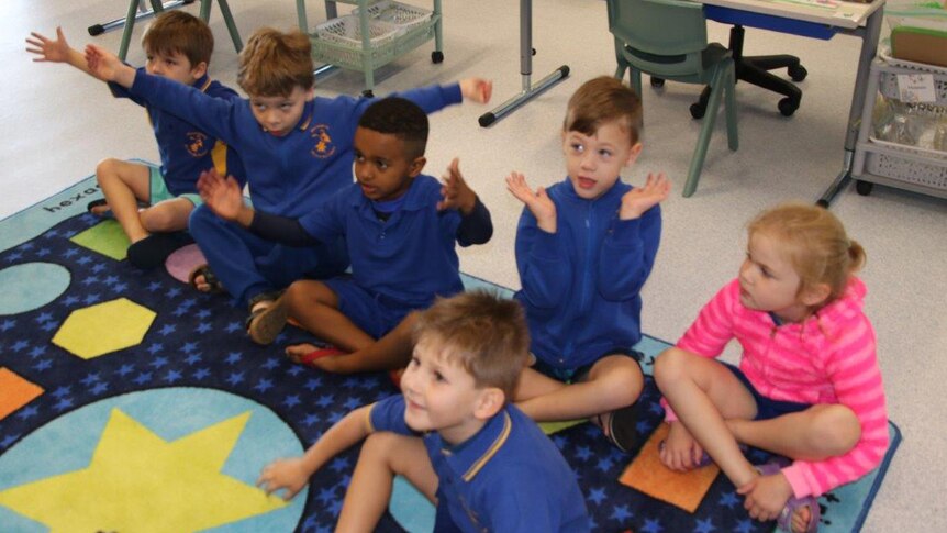 A group of primary school children in blue jumpers sit on a classroom floor.