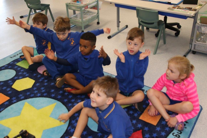 A group of primary school children in blue jumpers sit on a classroom floor.