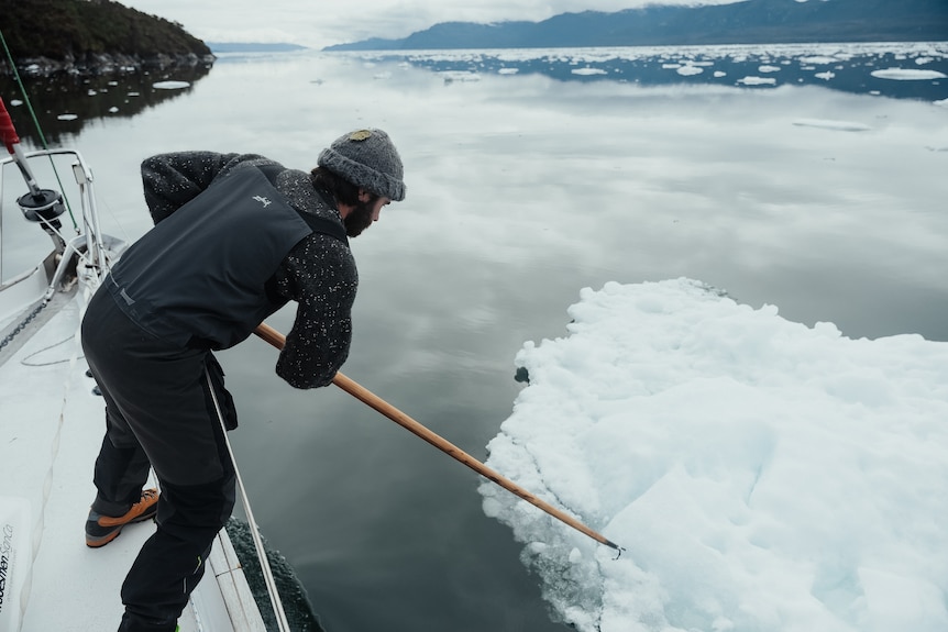 A man leans over the side of small boat an pushes away an iceberg with a pole.