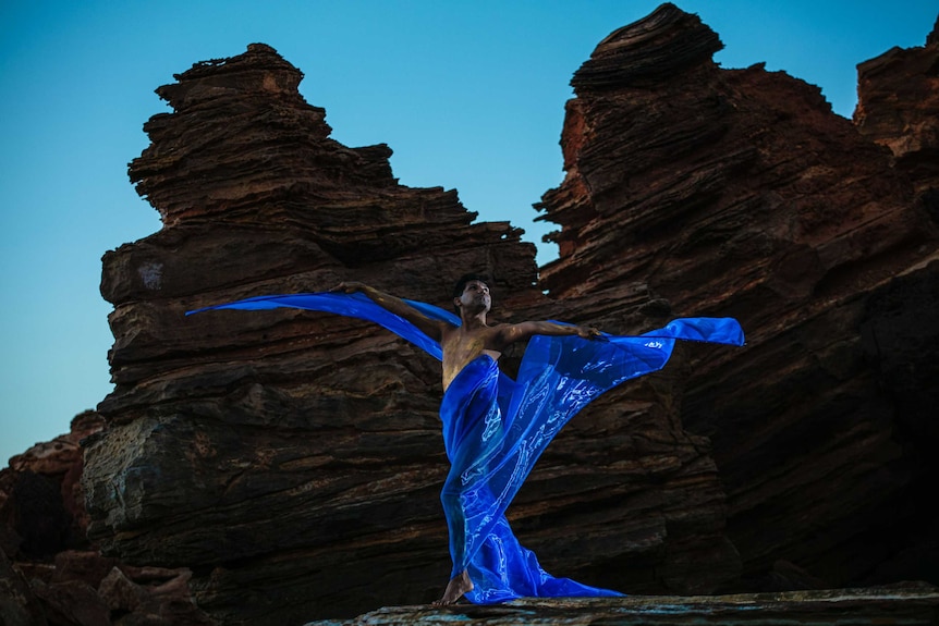 Man with blue ribbons flustered into wings, in front of large red rocks.