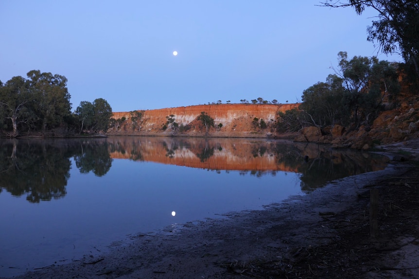 the sunrise above a river and red cliffs with the moon still in the sky
