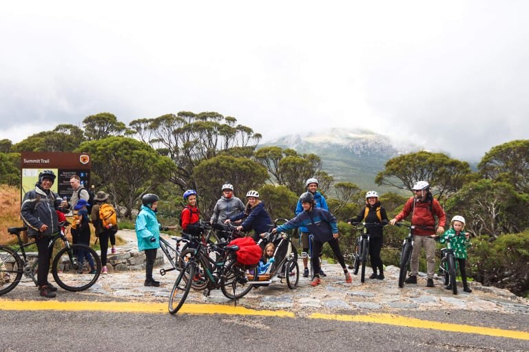 A group of adults and children stand with mountain bikes and unicycles on a mountain road.