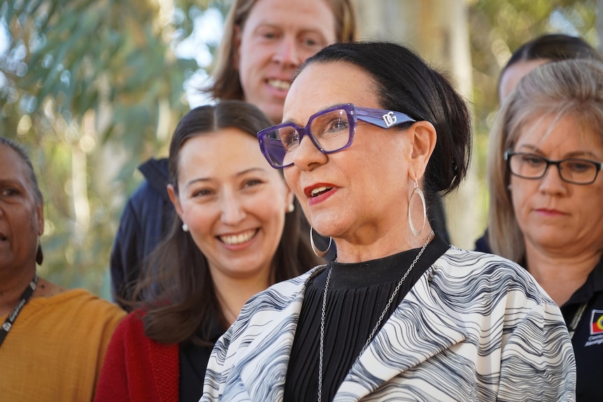 Linda Burney, standing in front of a group of people, holds a press conference outside with gum trees behind her