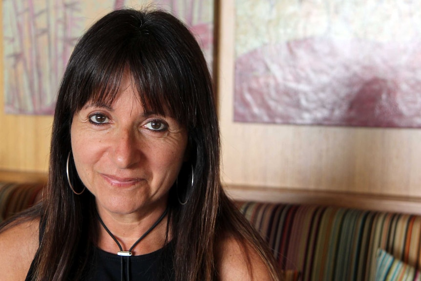 A woman with dark hair smiles into the camera against a wooden wall featuring paintings.