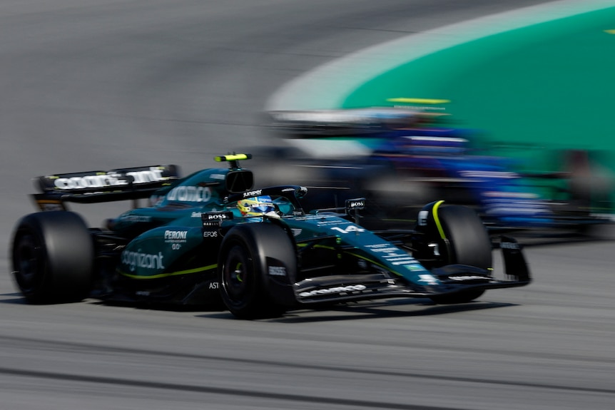 A green F1 car goes past a blue F1 car around a corner.