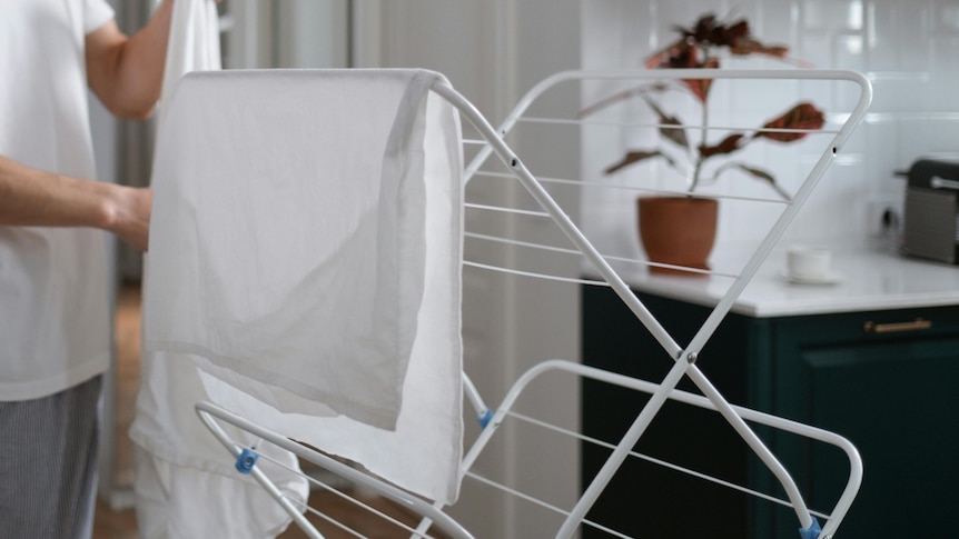 Close-up of someone hanging out white linen on a white indoor clothes drying rack located near a kitchen.