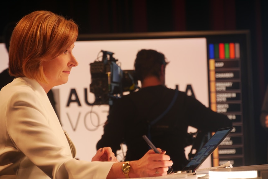 Woman wearing white jacket sitting at desk in studio with cameraman in background filming TV screen results.