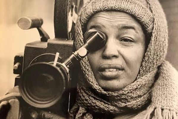 Black and white image of Tressie Sounders with scarf around her head, holding video camera and looking through viewfinder.