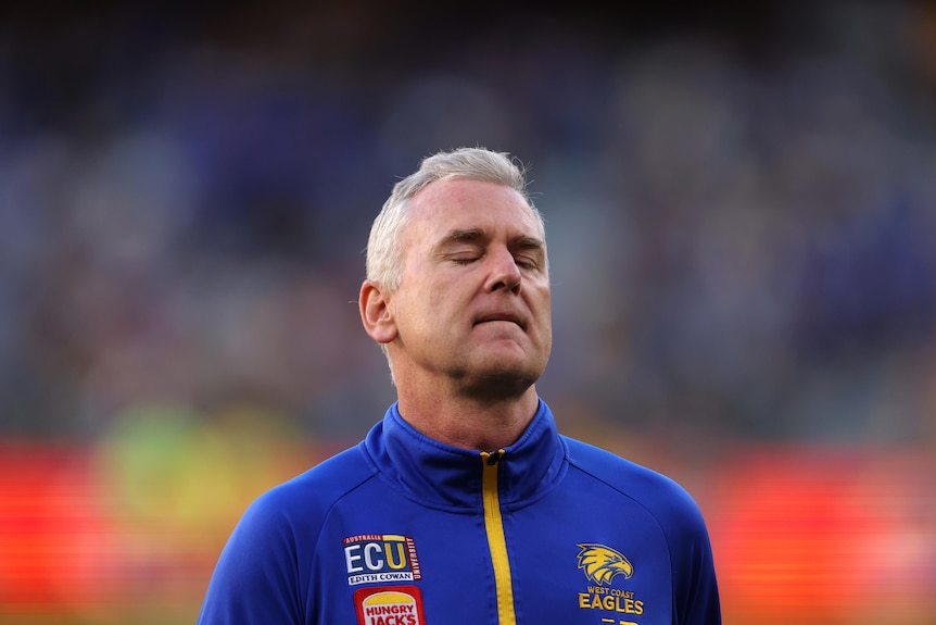 A man in a blue football club tracksuit looks exasperated with his eyes closed.