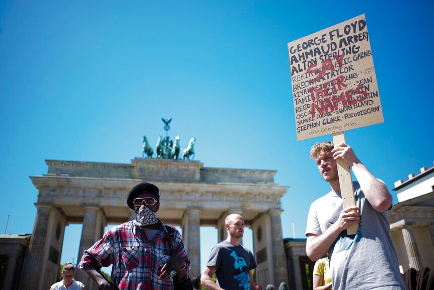 On a bright day, you view a person in front of the Brandenberg Gate holding a sign with the names of African Americans killed.