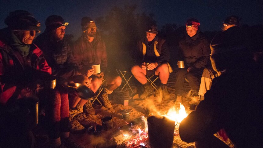 A group of people sit around a campfire in the dark.