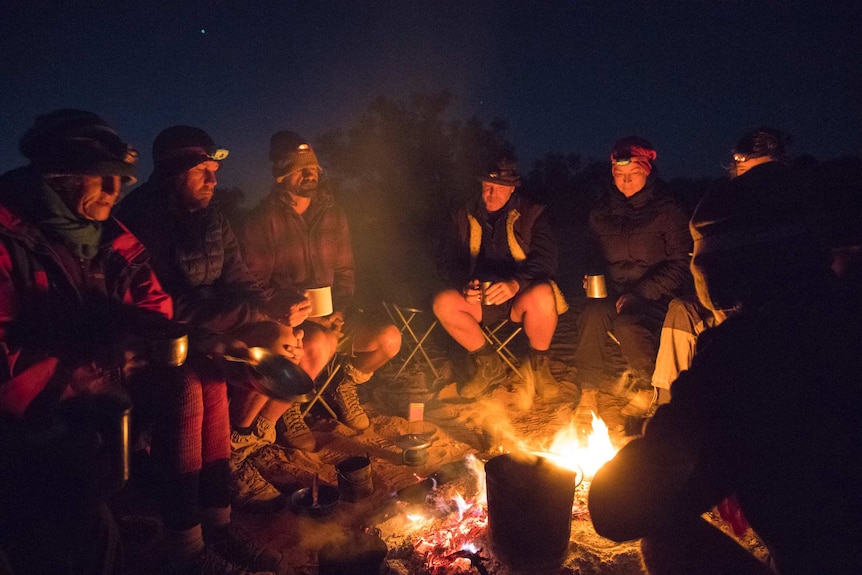 A group of people sit around a campfire in the dark.