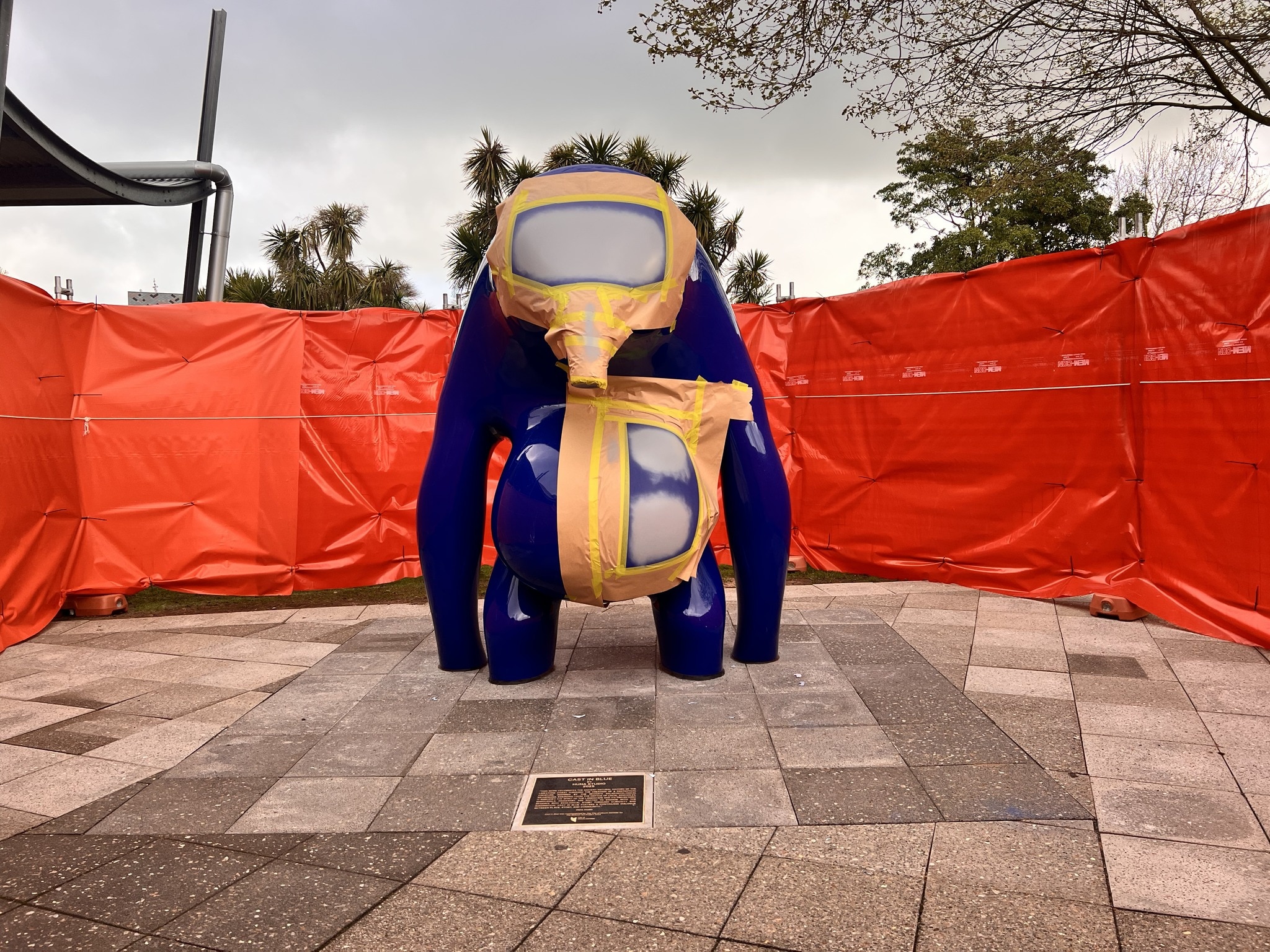 A large blue sculpture with gold tape and grey markings on it in front of an orange fence.