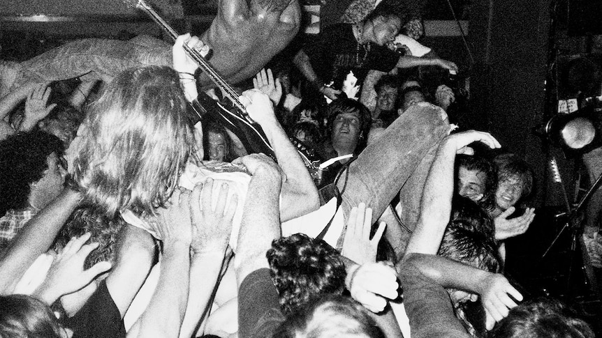 black and white photo of a man crowdsurfing while playing a guitar. other crowd members crowdsurf as well.