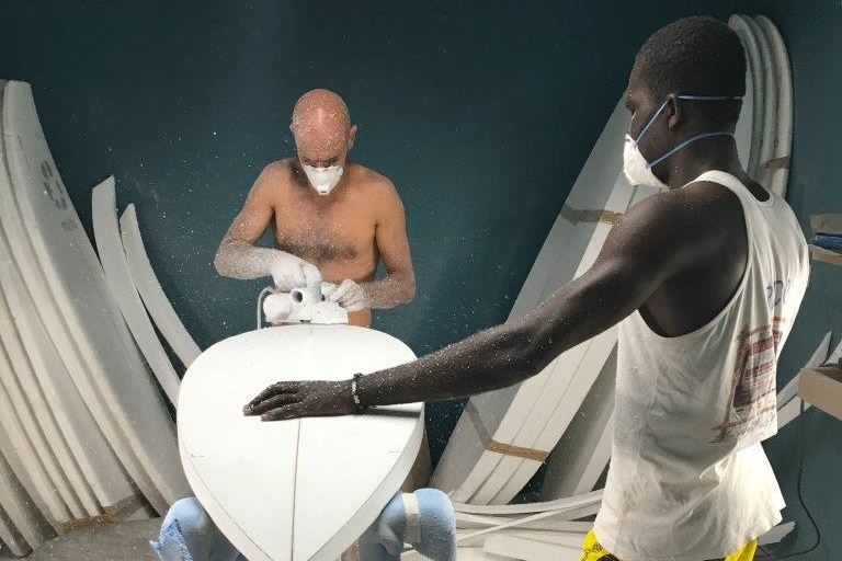 Paul Abbas wears a mask and sands a board as a local student watches