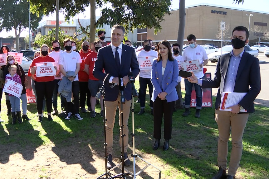 A man wearing a blazer standing with a woman also with a blazer in front of a group with signs