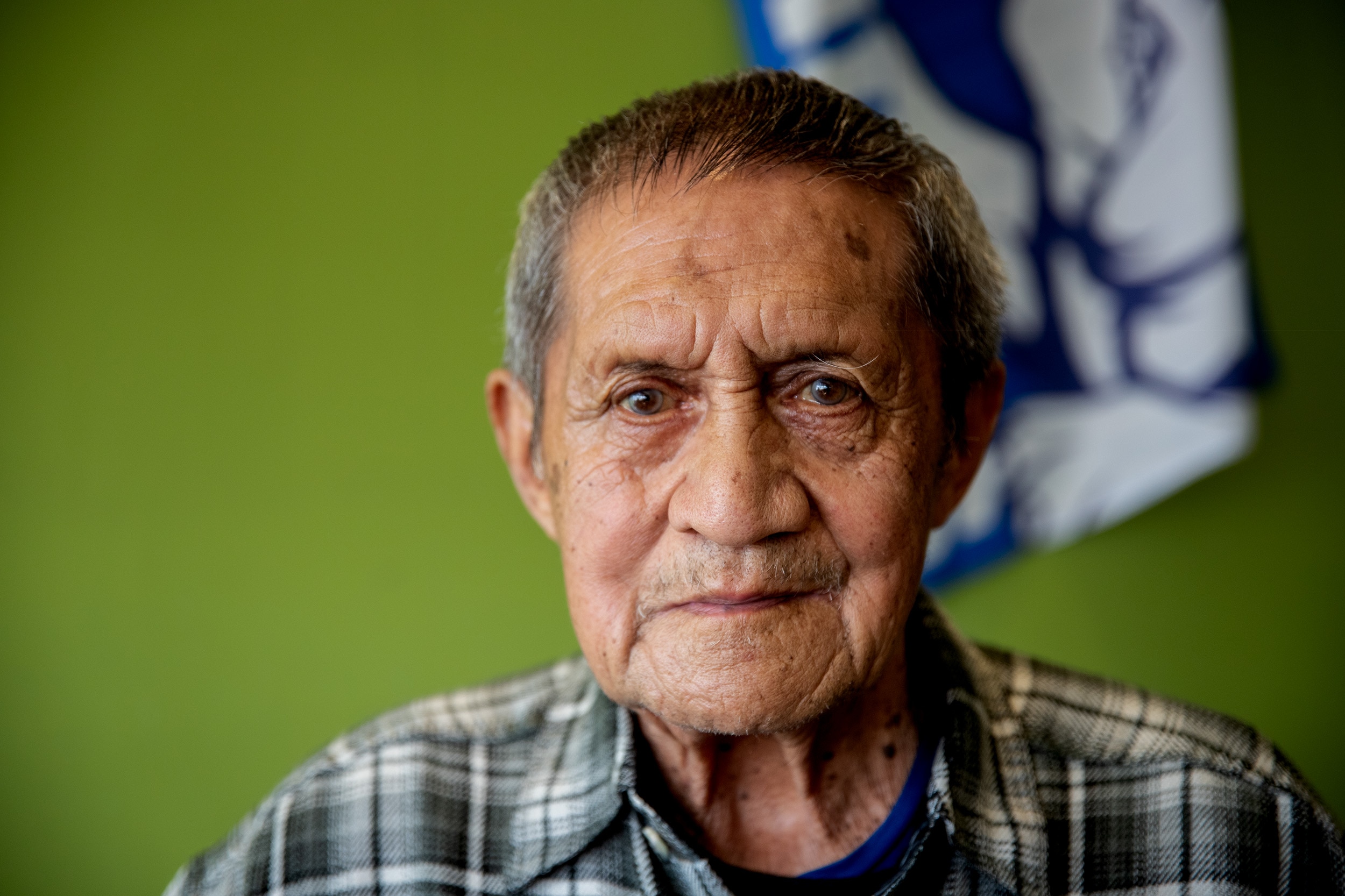 An older man wearing a flannel shirt stares in front of a green wall with a western bulldogs flag behind him