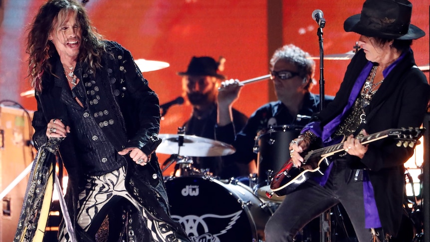 Aerosmith on stage. Steven Tyler dancing, Joe Perry playing the guitar, two others behind on the drums.