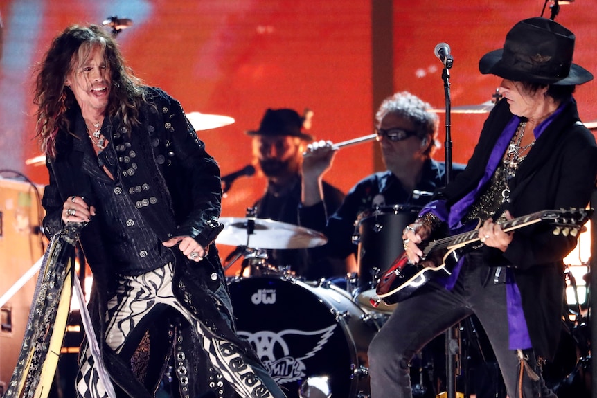 Aerosmith on stage. Steven Tyler dancing, Joe Perry playing the guitar, two others behind on the drums.