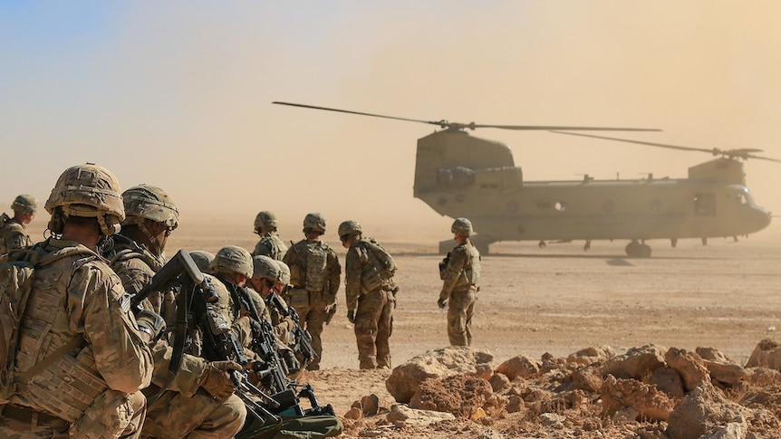 American soldiers in camouflage are seen from behind in a line in the desert while a helicopter blows up dust.