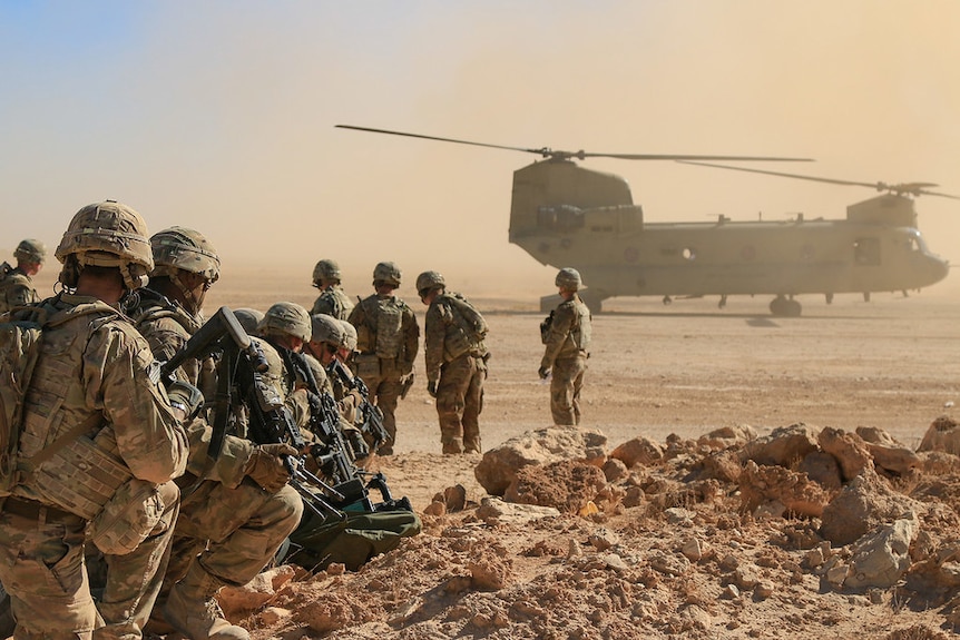 American soldiers in camouflage are seen from behind in a line in the desert while a helicopter blows up dust.