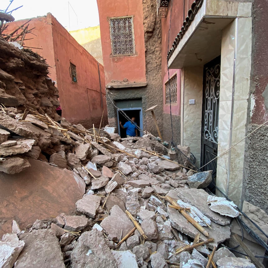 A man stands in the doorway of a collapsed building in front of him, with rubble everywhere