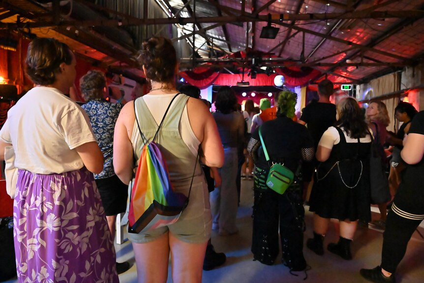 A group of people turn their back to the camera and face the stage. One person is wearing a rainbow backpack.