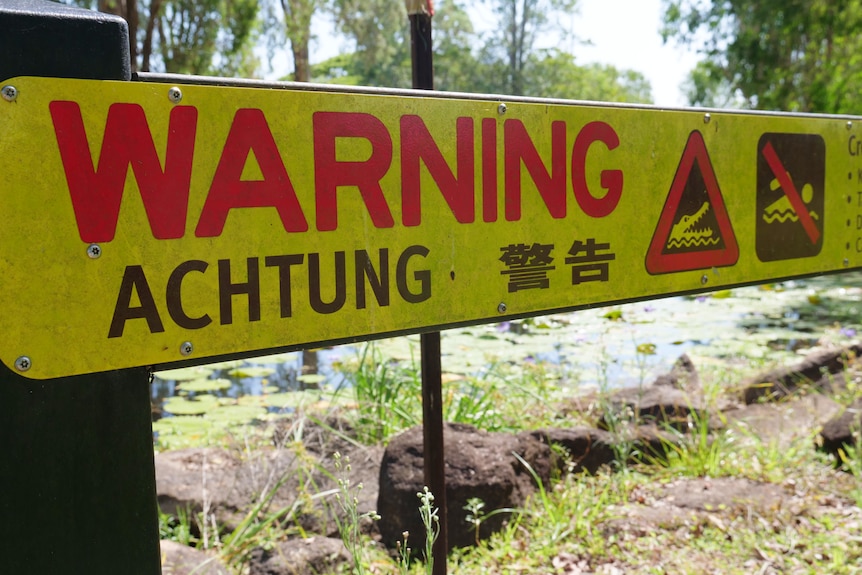 A yellow crocodile warning sign at a waterway, saying warning, achtung and signs for no swimming.