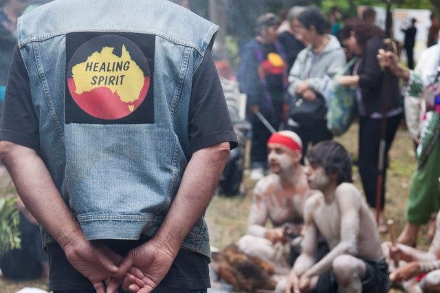 A man with his back to the camera stands and look at the Aboriginal male dancers performing a smoking ceremony.