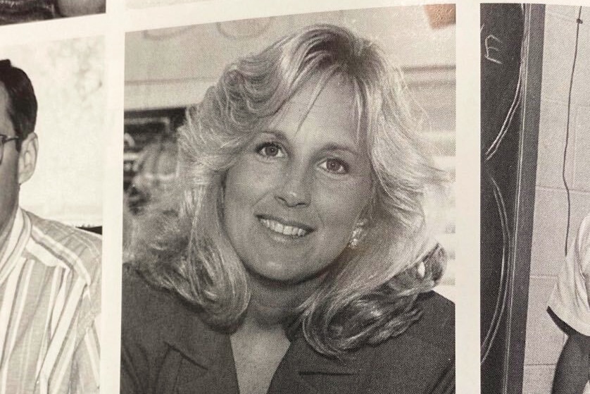 A woman with blond hair and wearing a blazer with a brooch smiles at the camera