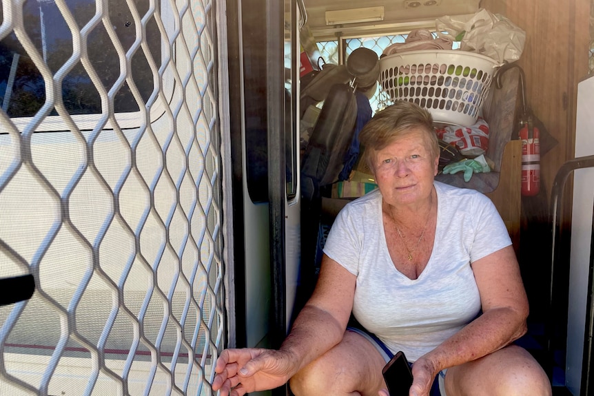 A woman in a white t-shirt sits next to a screen door with items including a washing basket piled behind her.