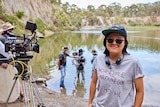 A Taiwanese woman with headphones around her neck and hat and sunglasses stands in front of a muddy lake, film cameras and crew