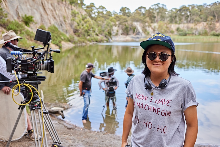 A Taiwanese woman with headphones around her neck and hat and sunglasses stands in front of a muddy lake, film cameras and crew