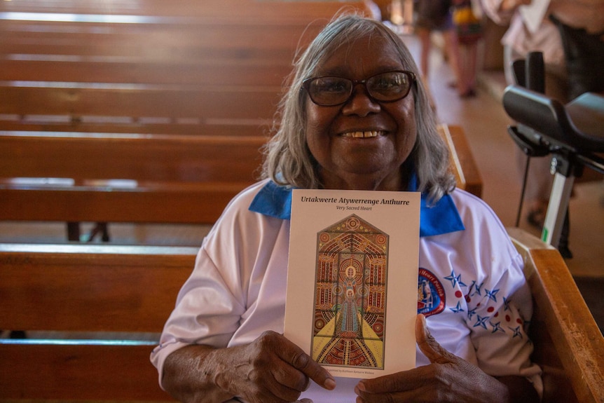 An Aboriginal woman sits in a pew, smiling, and holding a brochure with an artwork on the cover.