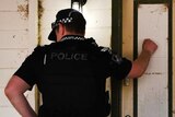 A policeman knocks on a front door while his partner checks a clipboard