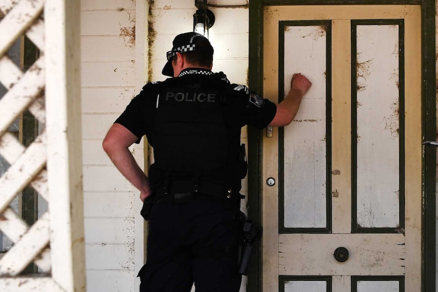 A policeman knocks on a front door while his partner checks a clipboard