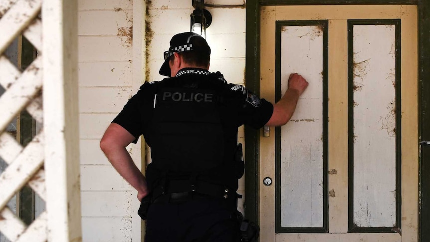 A policeman knocks on a front door while his partner checks a clipboard