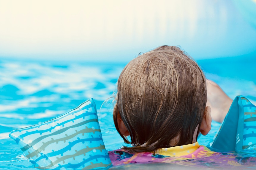 Back of a young child's head. Child wears two arm-floaties and clear blue pool water can be seen in background.