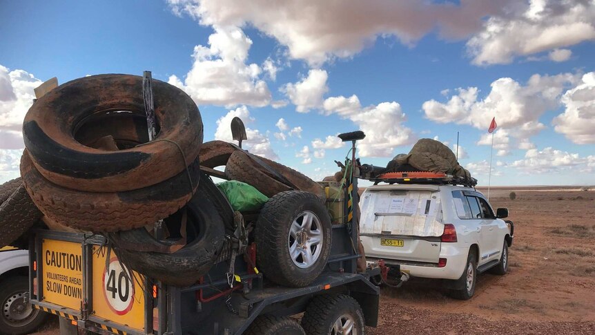 A white four-wheel-drive connected to a trailer filled with tyres and waste has stopped on the red dirt.