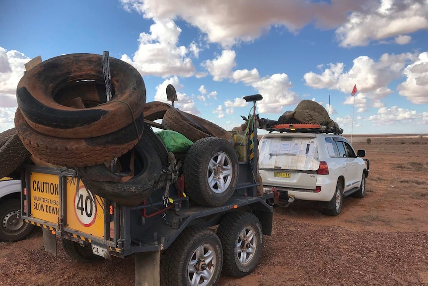 A white four-wheel-drive connected to a trailer filled with tyres and waste has stopped on the red dirt.