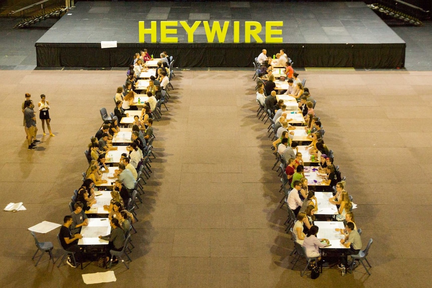 Two long rows of tables with young people sitting across from each other in front of large Heywire sign.
