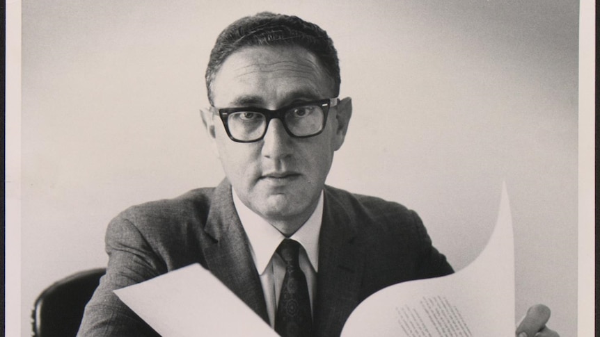 A young henry kissinger in glasses and in front of books 