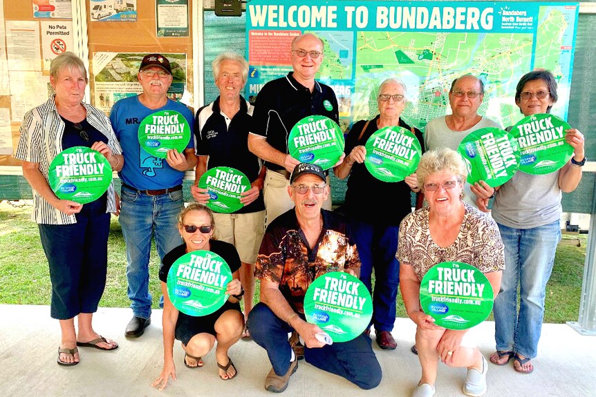 A group of campervanners hold out "I'm truck friendly" stickers in front of a welcome to Bundaberg sign.