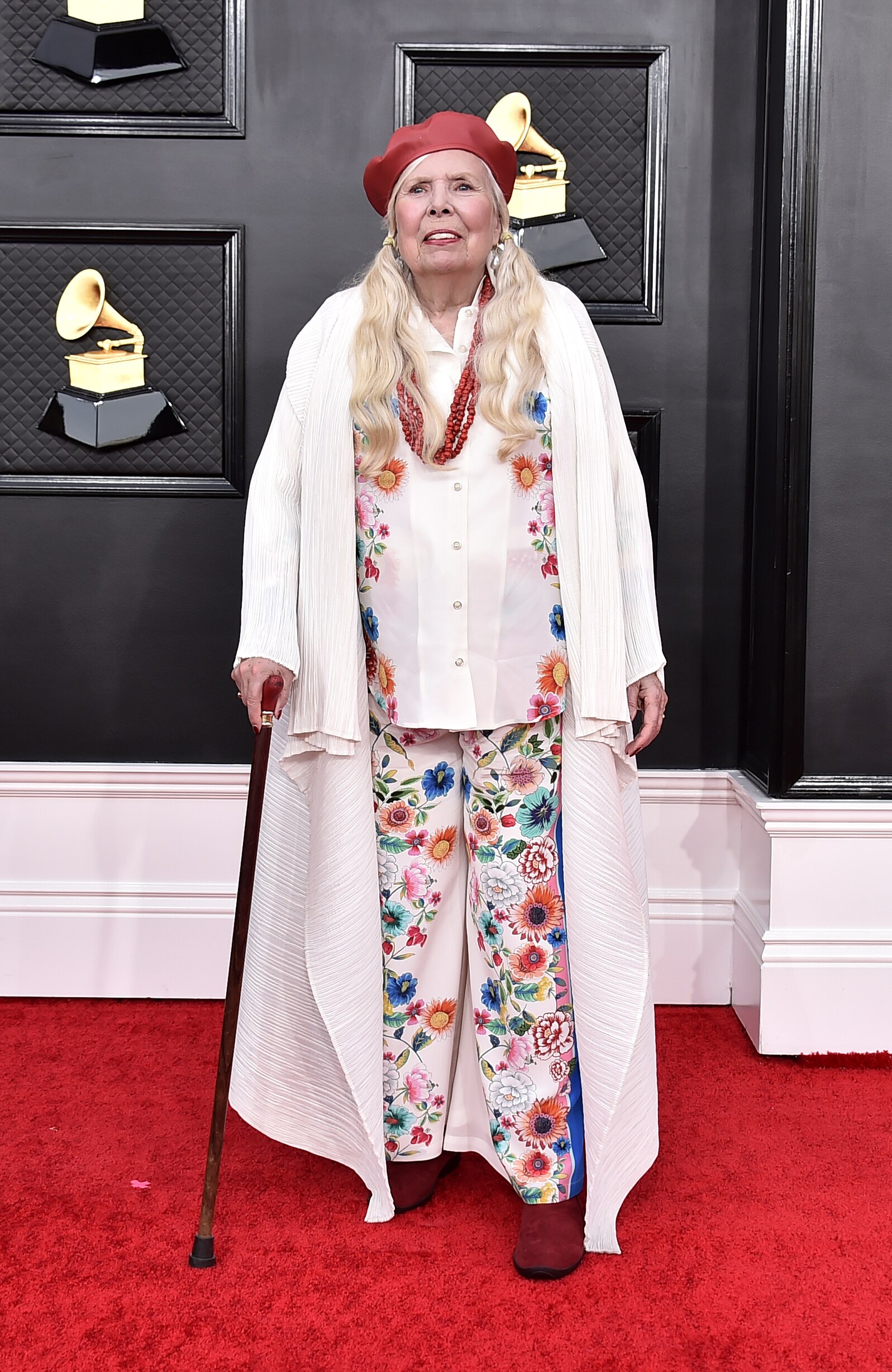 artist joni mitchell smiles on the grammys red carpet wearing a red beret and a white floral outfit holding a walking stick