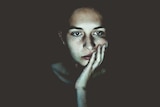 A woman sitting in a dark room stares blankly at a screen, which illuminates her face.