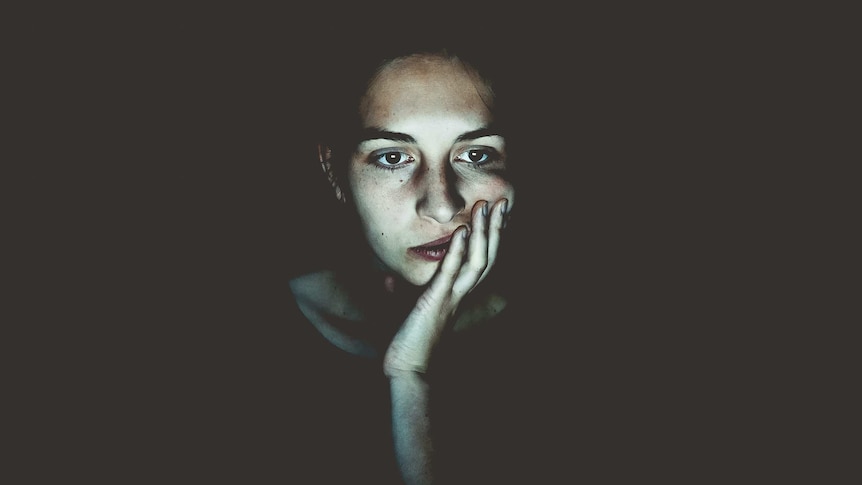 A woman sitting in a dark room stares blankly at a screen, which illuminates her face.
